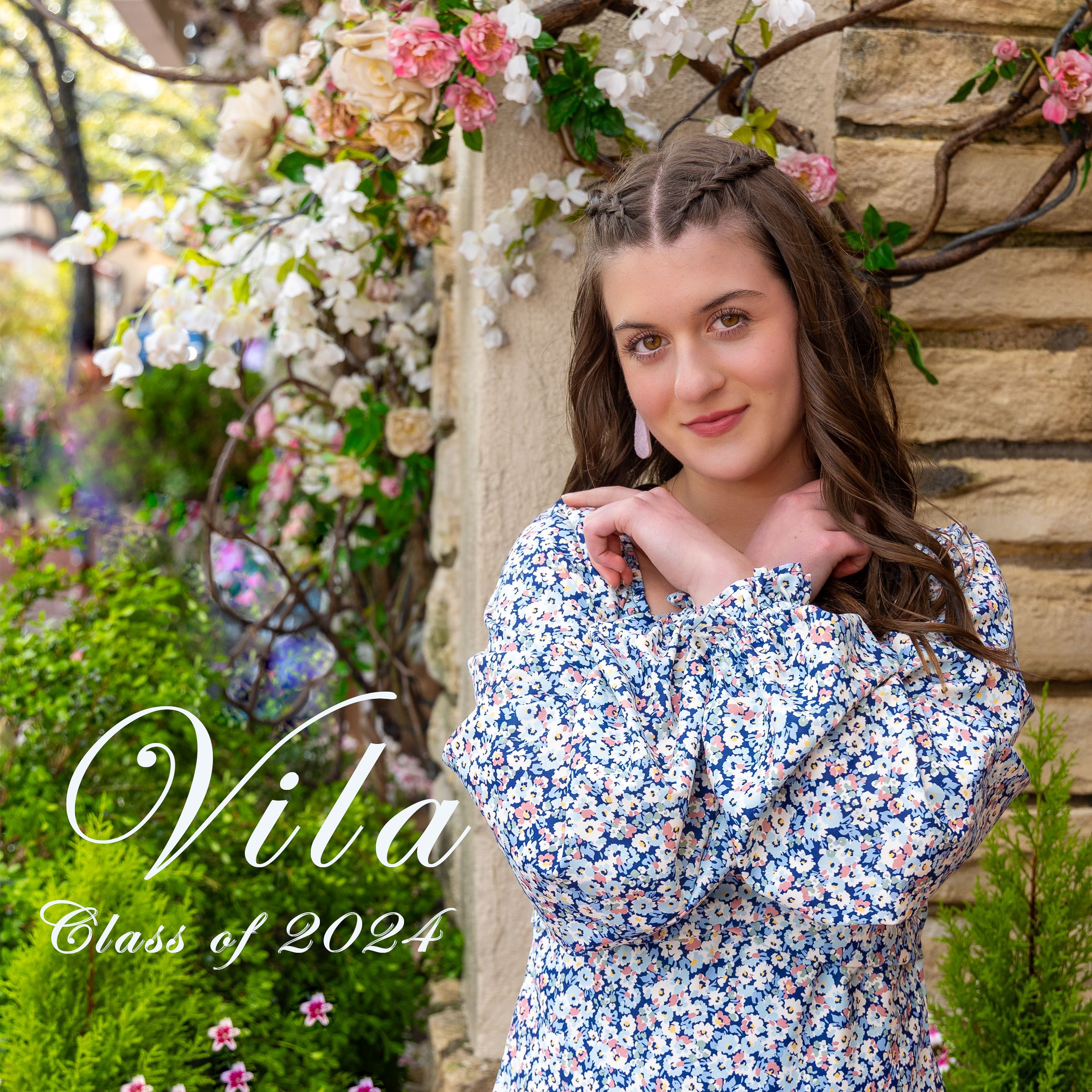 Spring graduation portrait with blue and white floral dress against stone wall with blooming pink and white flowers.