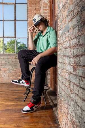 Person in green polo and black pants sitting on barstool near exposed brick wall and window. senior photos