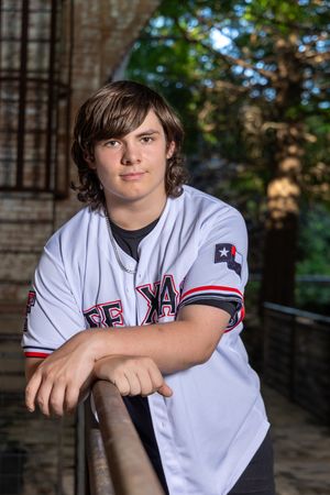 Young athlete wearing Texas baseball jersey poses against rustic brick wall background.