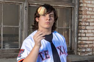 A baseball player in a white and blue uniform stands thoughtfully with their hand near their face.