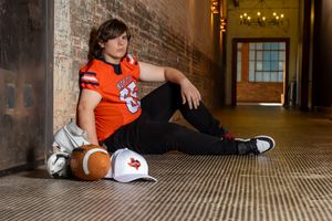 Young person in orange sports jersey sits on tiled floor with helmet and sneakers in dimly lit corridor. senior photos