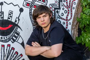A young person in a black t-shirt poses with crossed arms against a wall with artistic graffiti and murals.