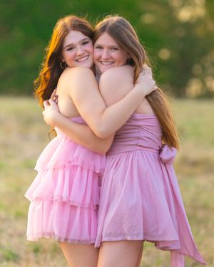 Two people in pink dresses embrace and smile while posing outdoors with a blurred natural background.