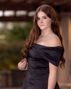 A person with brown hair poses in a black off-shoulder dress against a blurred background.