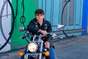 A motorcyclist in dark clothing sits on a parked motorcycle against a blue and green painted wall background.