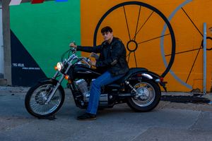 A motorcycle rider sits on a parked black cruiser bike against a colorful wall with geometric shapes.