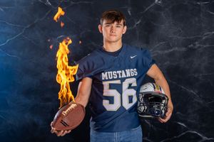 Football player in navy Mustangs jersey number 56 poses with helmet and ball against dark background with flame effects.