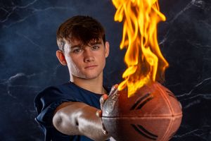 Basketball player stands against dark background with dramatic flames and lighting effects creating dynamic sports portrait.