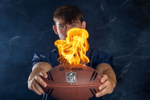 A flaming NFL football being held against a dark background with dramatic fire and smoke effects.