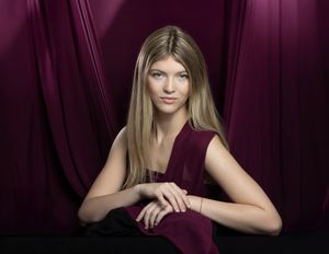 Young blonde woman in burgundy sleeveless top poses against matching draped fabric backdrop in professional portrait.