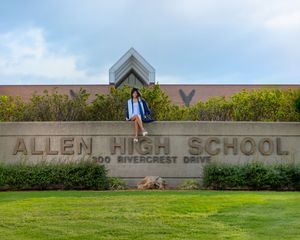 Student stands by Allen High School entrance sign surrounded by greenery and modern architectural features.