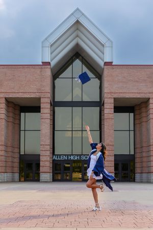 Graduate in blue cap and gown performs celebratory leap while tossing cap in front of brick high school entrance.