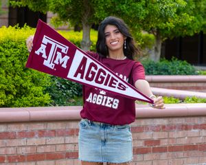 College student displays Texas A&M Aggies spirit banner while wearing maroon shirt in outdoor campus setting.