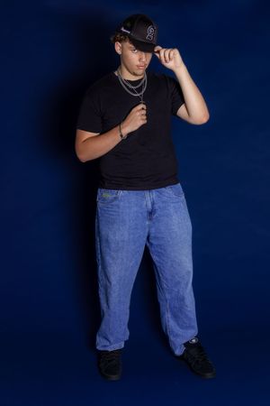 Person wearing a black baseball cap, black t-shirt and baggy blue jeans poses against dark blue background.