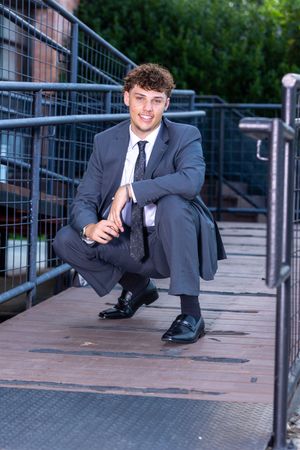 A person in a formal suit poses playfully on an outdoor deck with metal railings.