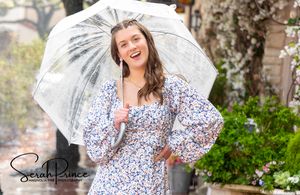 A woman in a floral dress poses with a white lace parasol in a garden setting during a photo session.