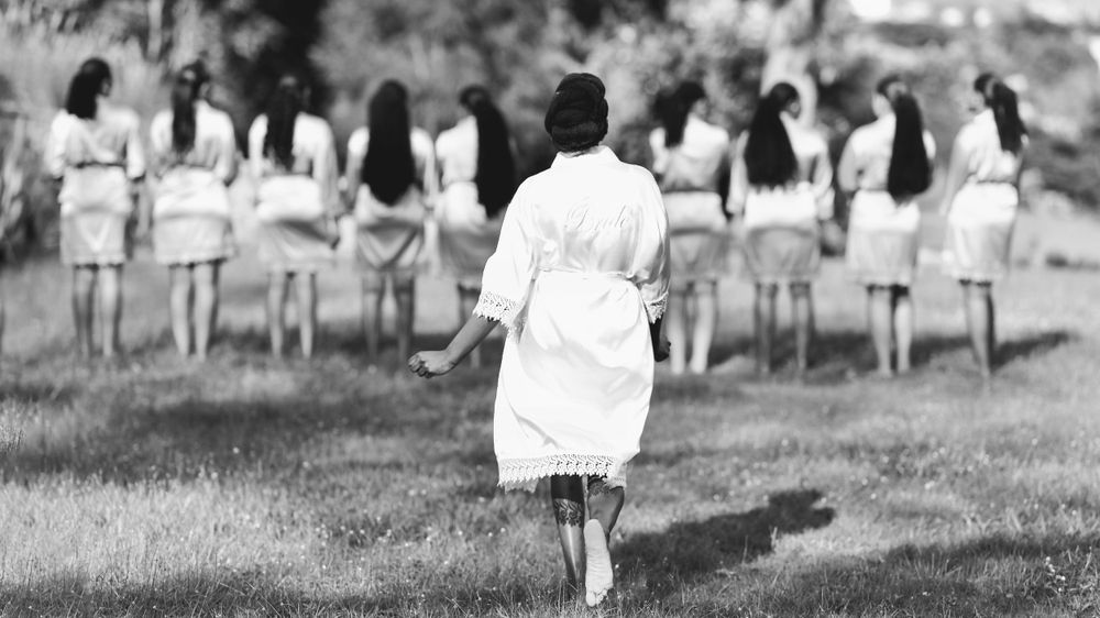 Black and white photo showing figure walking towards line of seated people outdoors.