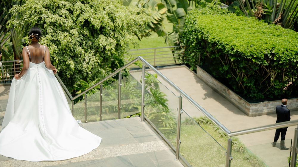 Wedding scene with bride in flowing white dress standing on outdoor stairs near lush greenery.