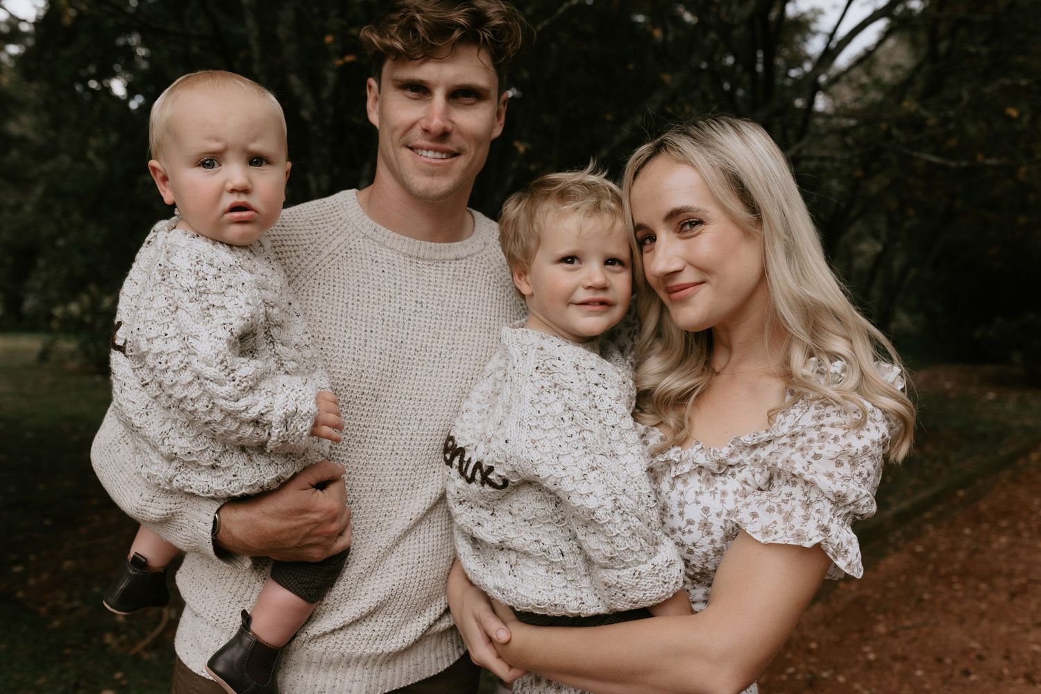 A family wearing coordinated cream knit sweaters poses together outdoors in a natural setting.