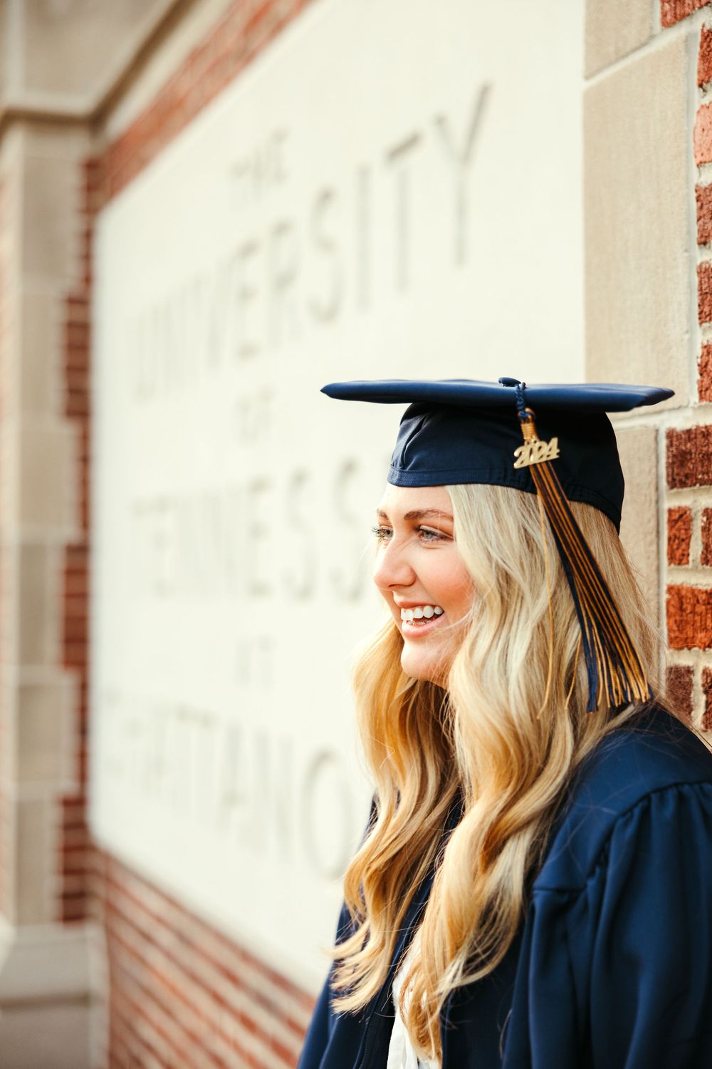 Graduate in blue cap and gown smiling against a brick wall during graduation photos.