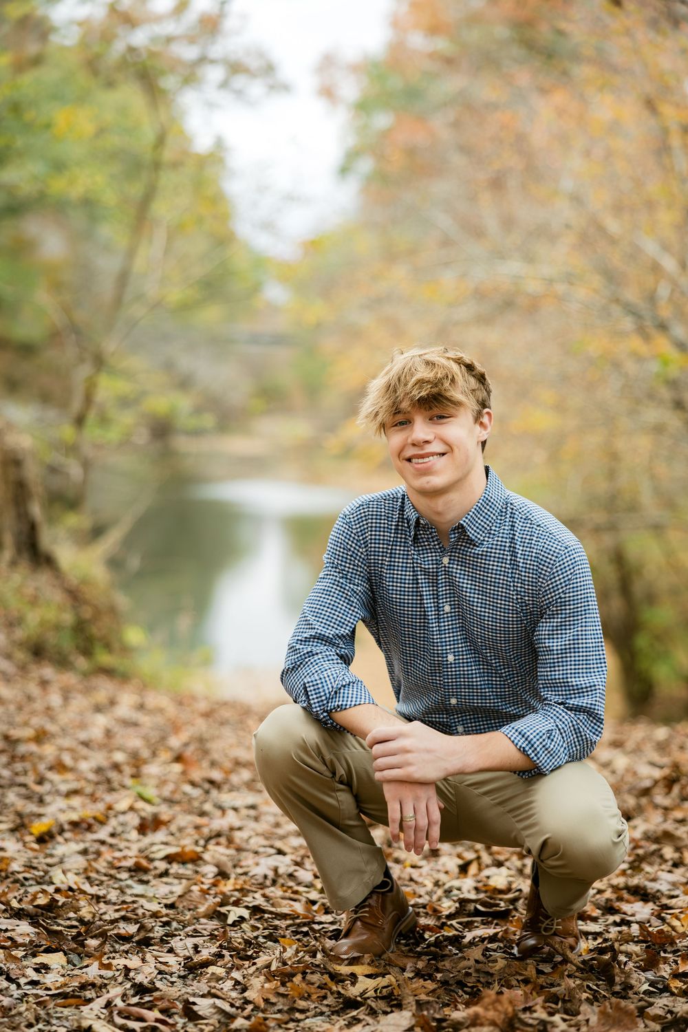 Autumn outdoor portrait with someone crouching on fallen leaves by a tranquil river with fall foliage in background.