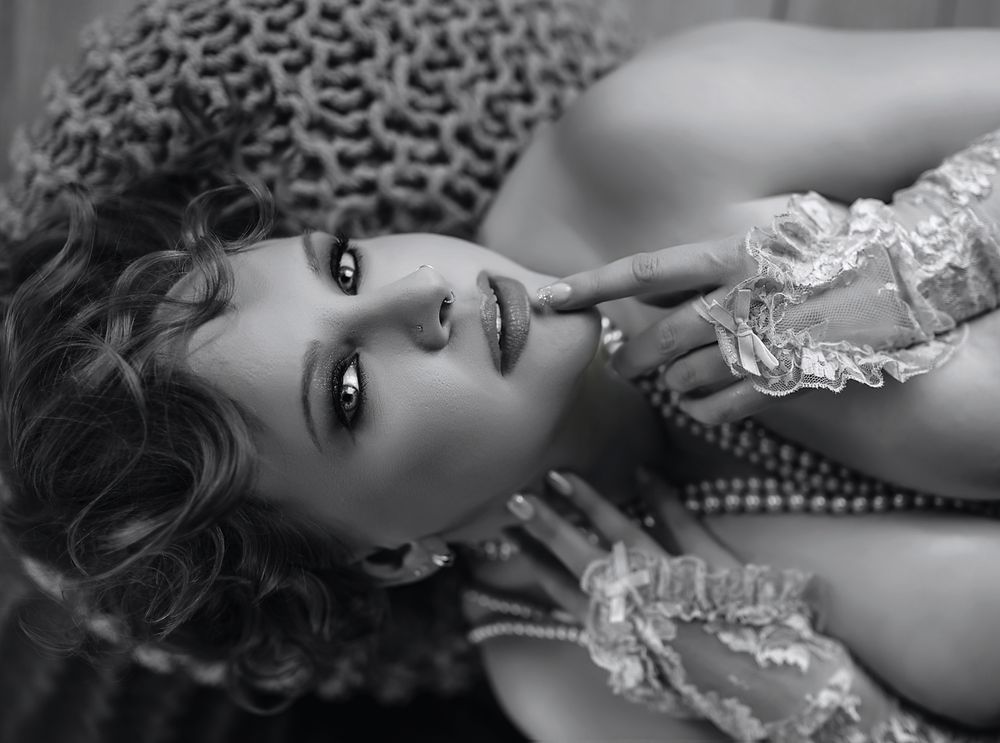 A black and white portrait of someone with curly hair and lace clothing lying down in an artistic pose.