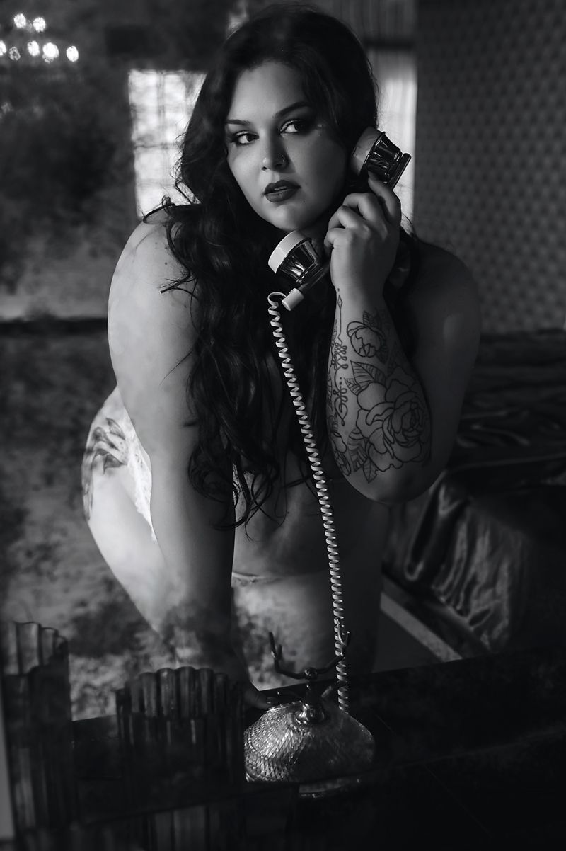 Black and white photo of a tattooed woman holding a vintage rotary phone receiver in a moody, dimly lit setting.
