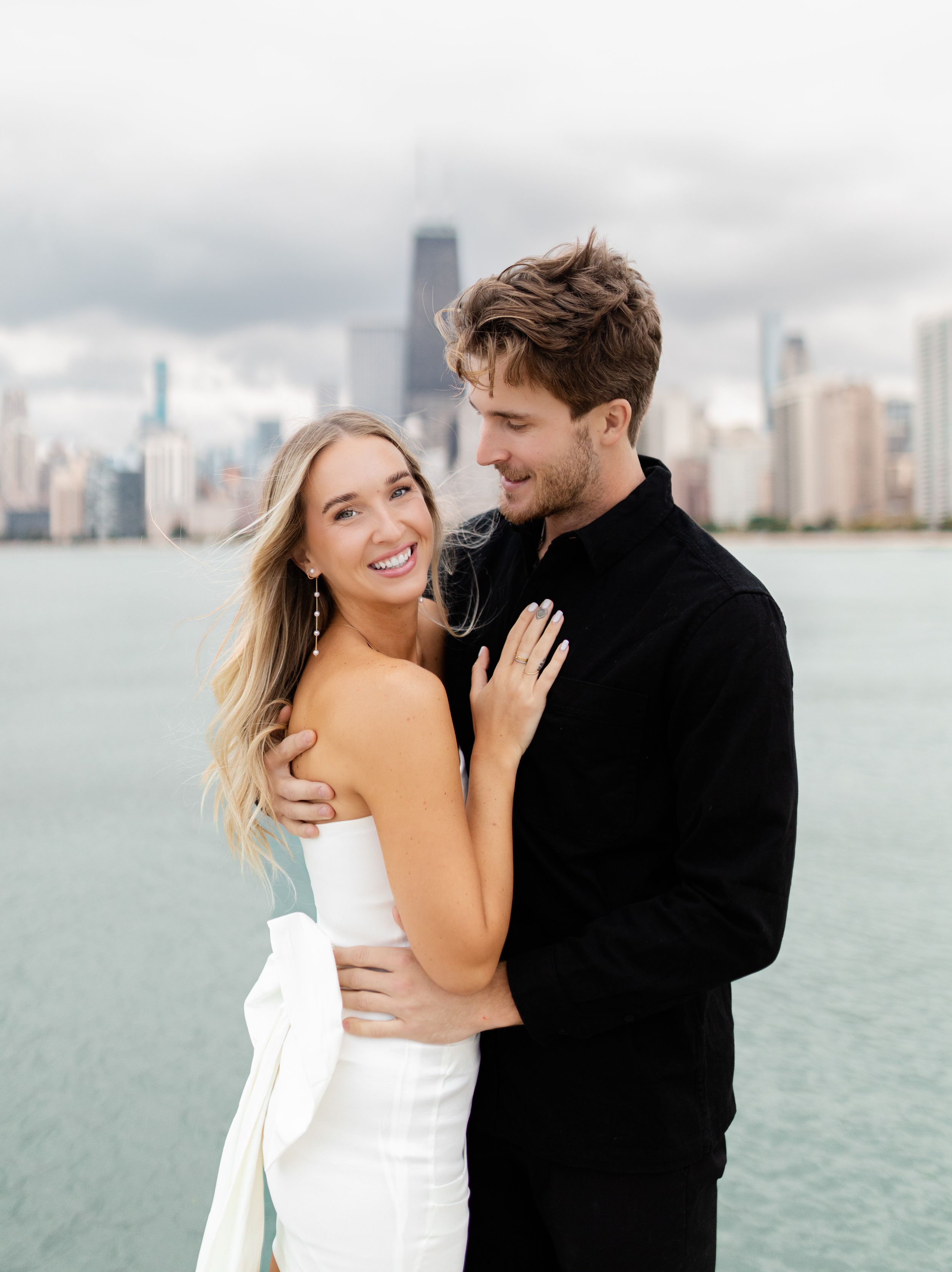 Couple embracing with Chicago skyline visible across Lake Michigan on cloudy day.