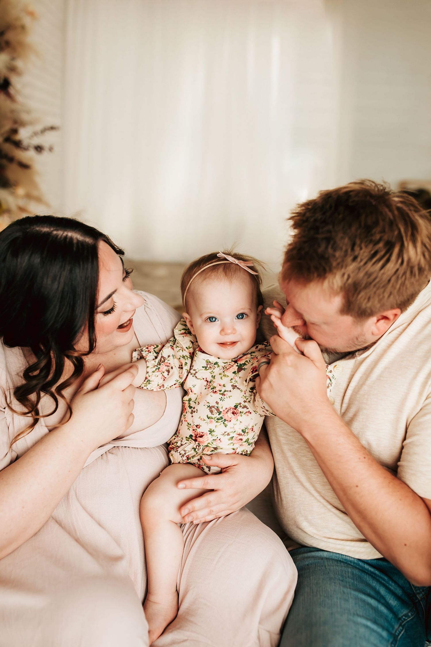 Sweet family moment captured in warm lighting as parents lovingly interact with their baby wearing a floral outfit.