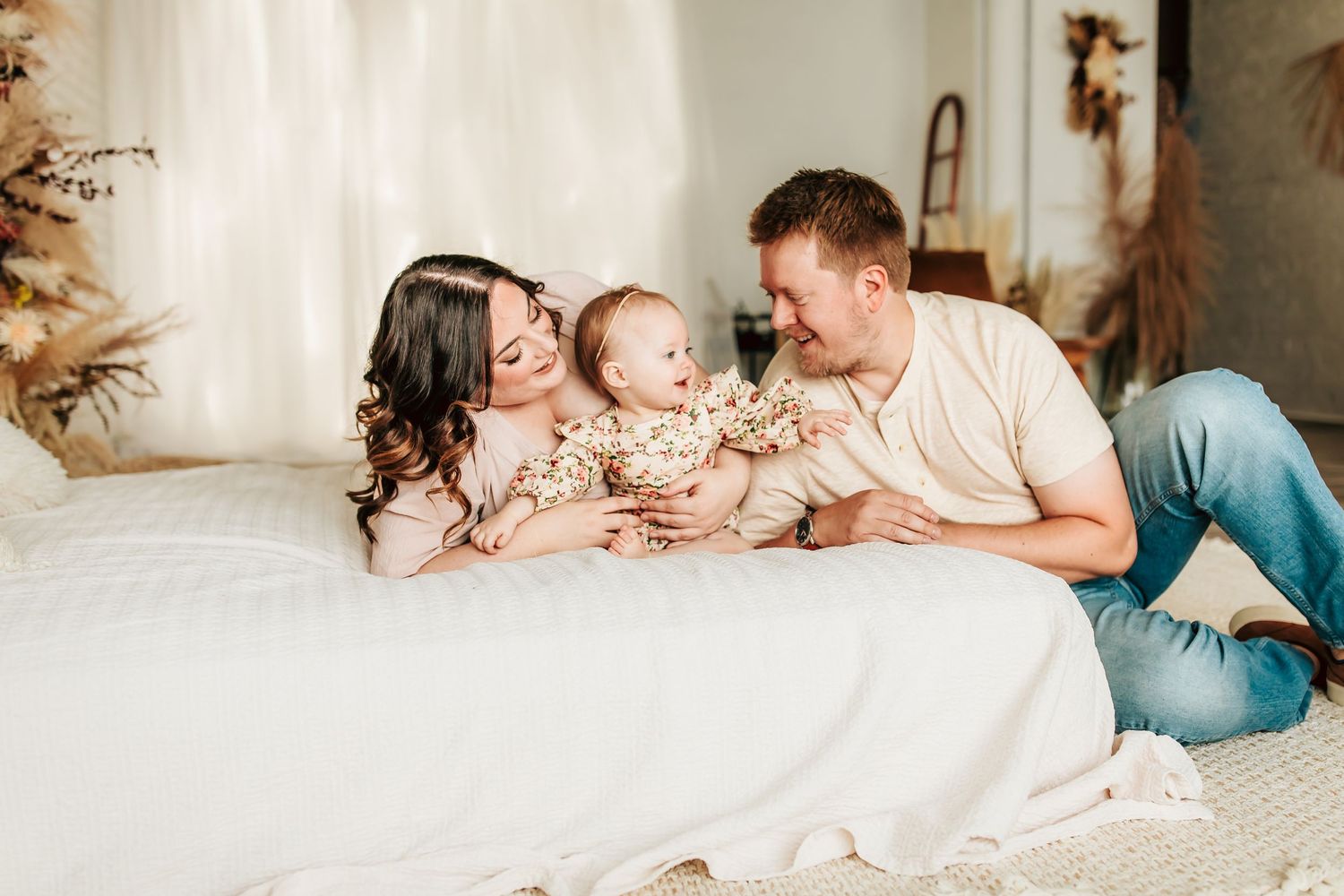 A family lies together on white bedding, sharing joyful moments with their baby in a light and airy setting.