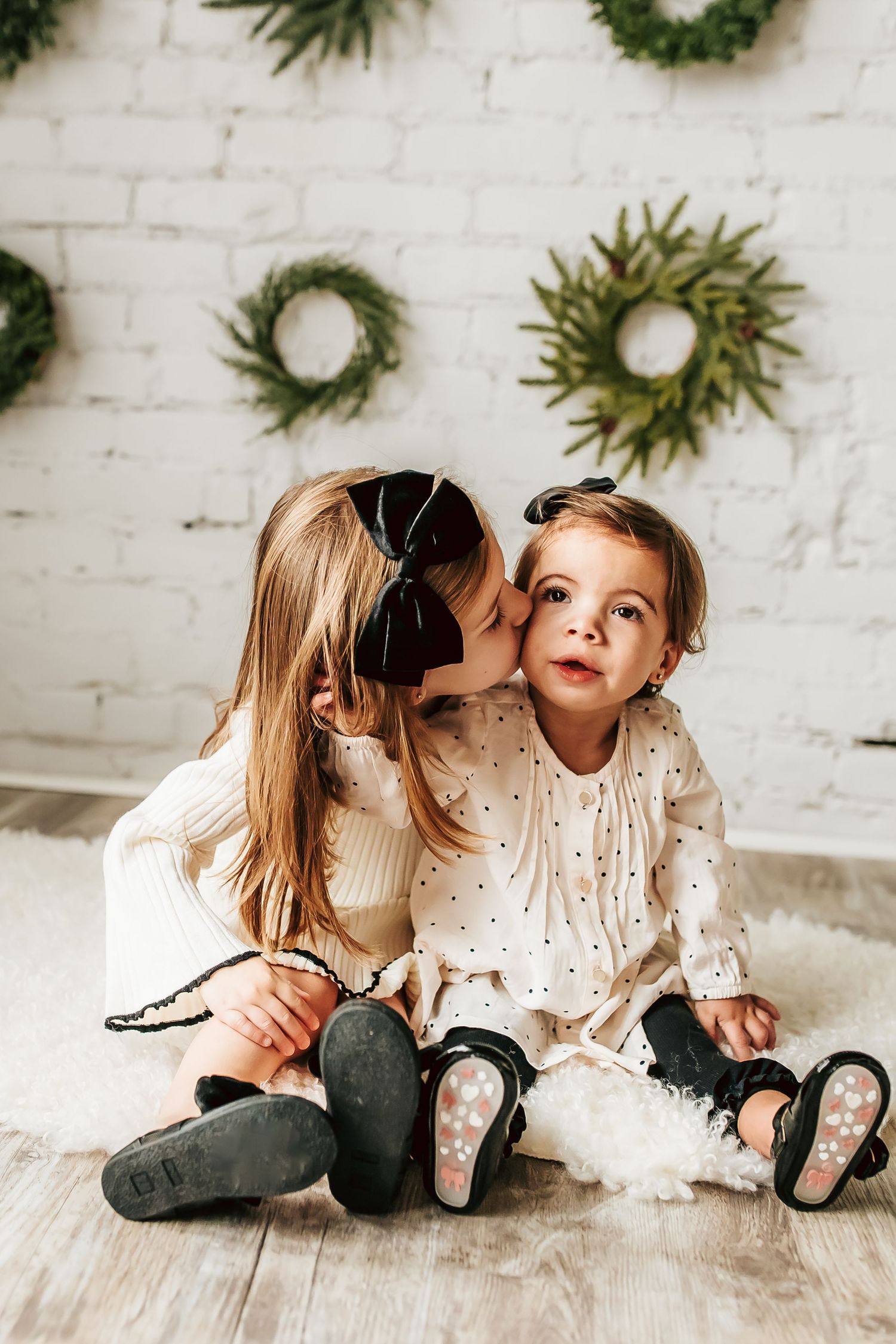 Two children in white dresses sit on wooden floor with evergreen wreaths decorating white brick wall behind them.