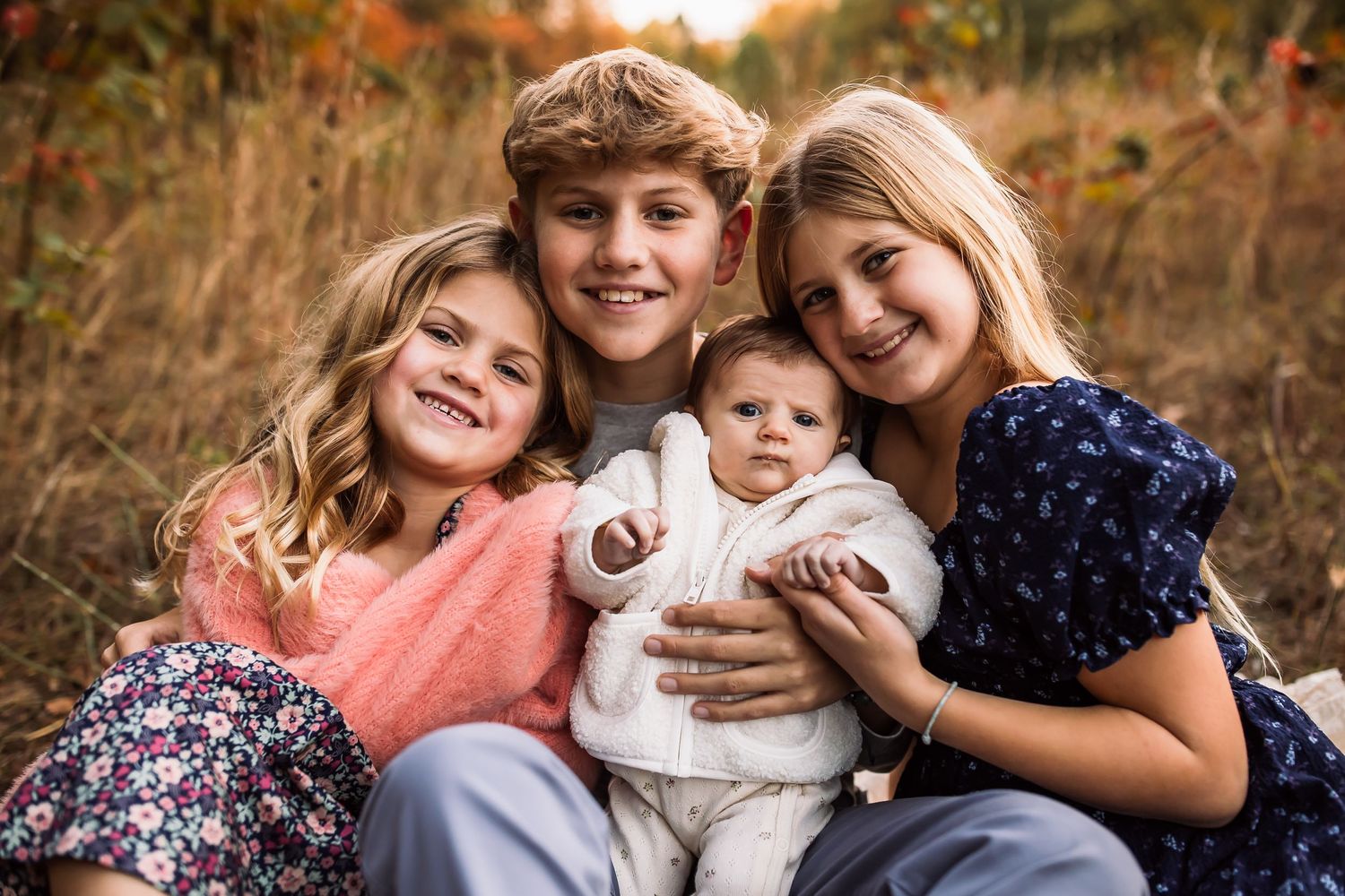 Four siblings sit together outdoors during autumn with fallen leaves and golden light in the background.