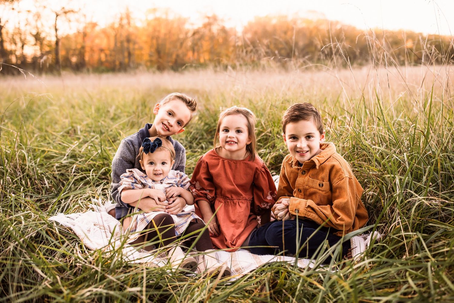 Children sit together on a blanket in a golden autumn field at sunset with tall grass surrounding them.