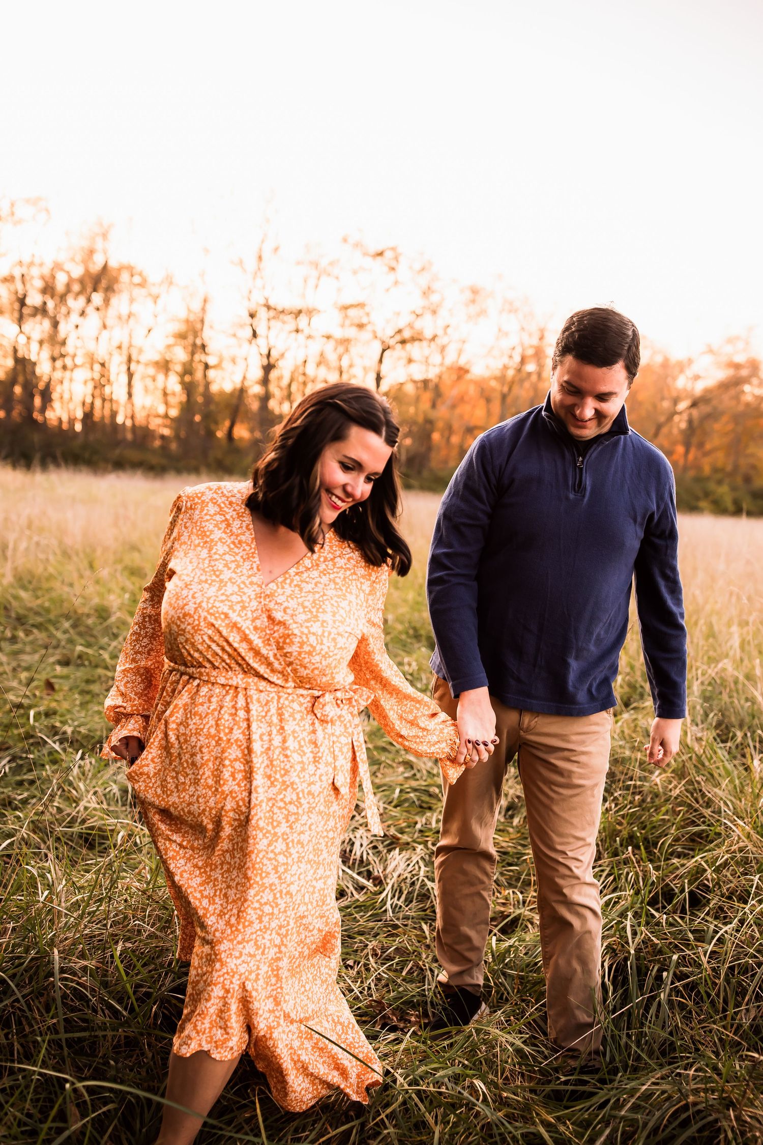 A couple walks together through a golden field at sunset, with her wearing a flowy yellow dress and him in a navy sweater.