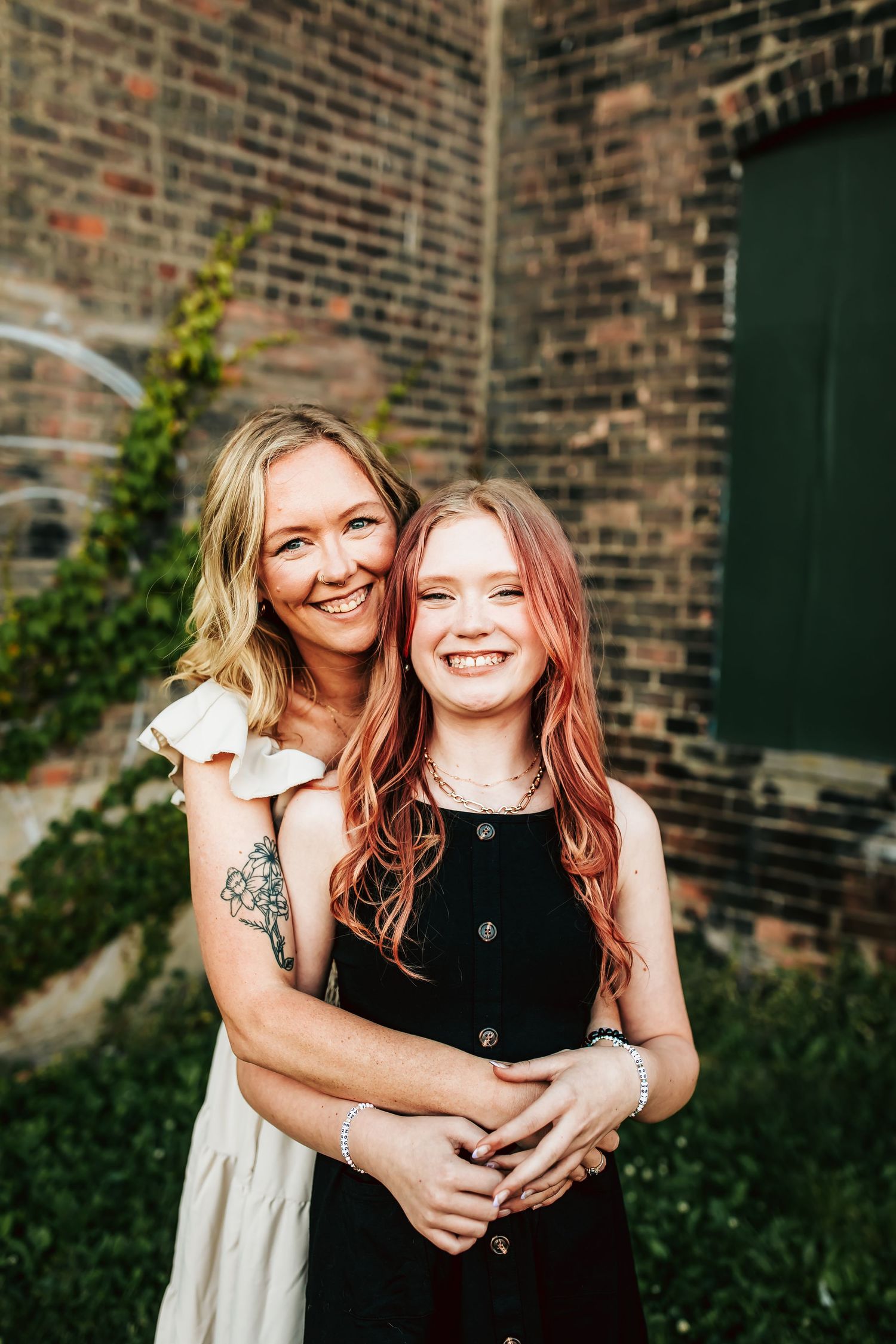 Mother and daughter embrace and laugh together while standing in front of a brick wall with climbing vines.