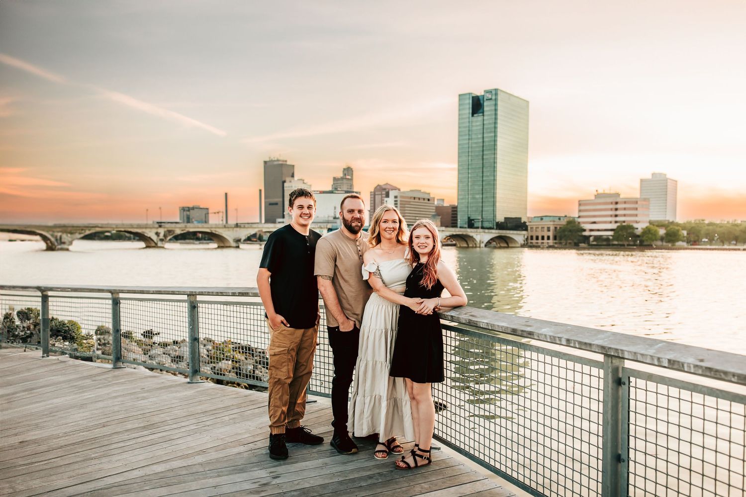 A family stands together on a waterfront boardwalk at sunset with Toledo's downtown skyline visible across the river.