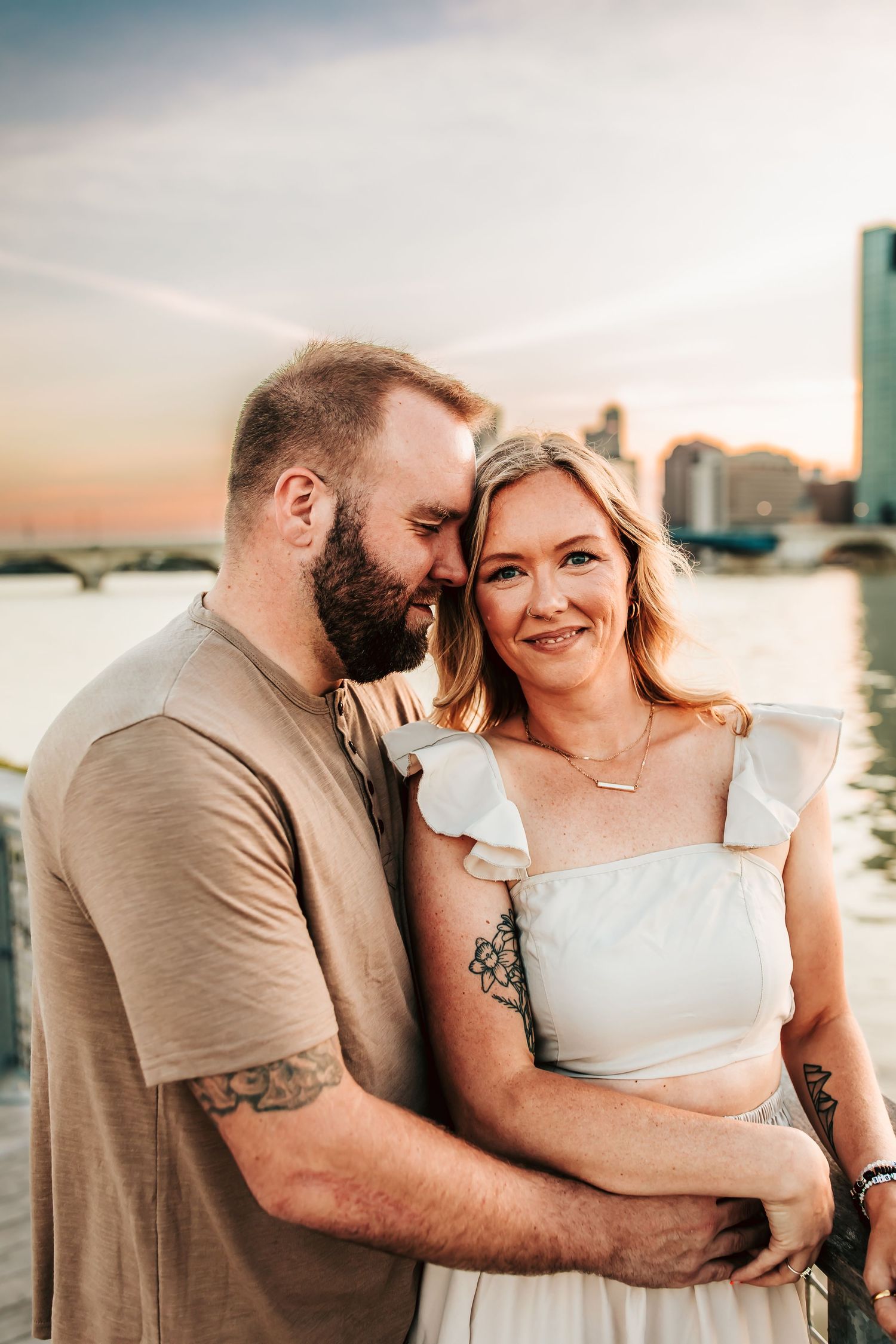 A couple shares an intimate moment at sunset along a waterfront with a city skyline in the background.
