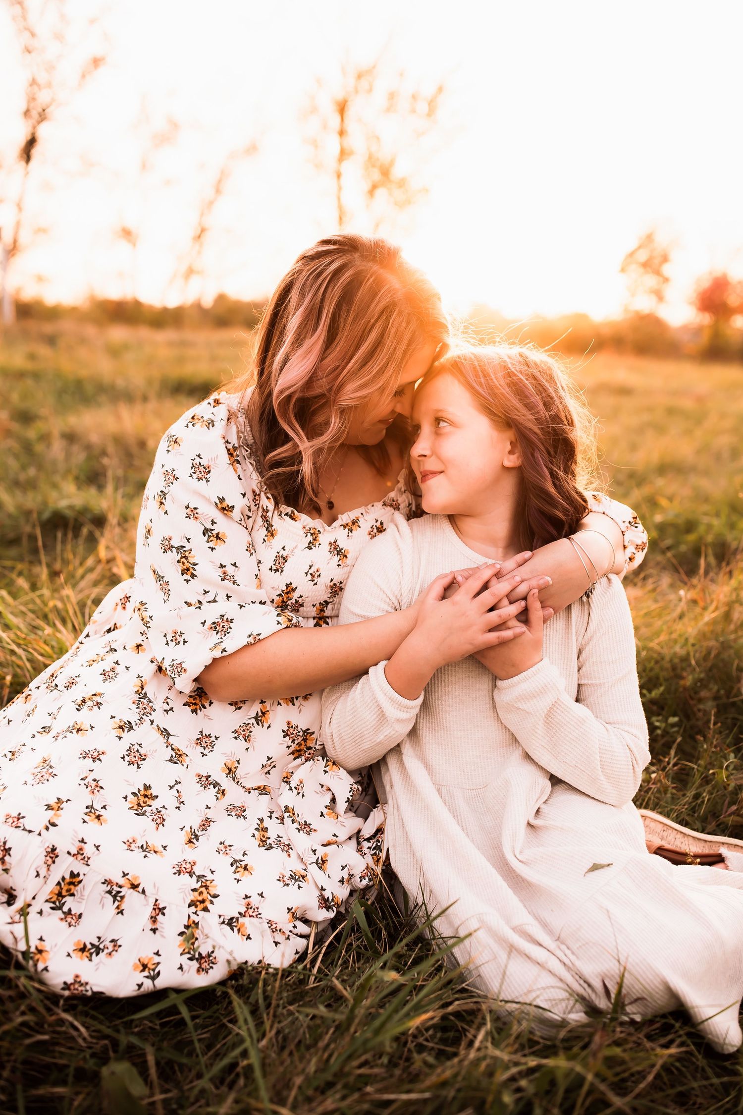 Two people in white dresses share a tender embrace while sitting in a sunlit meadow at golden hour.