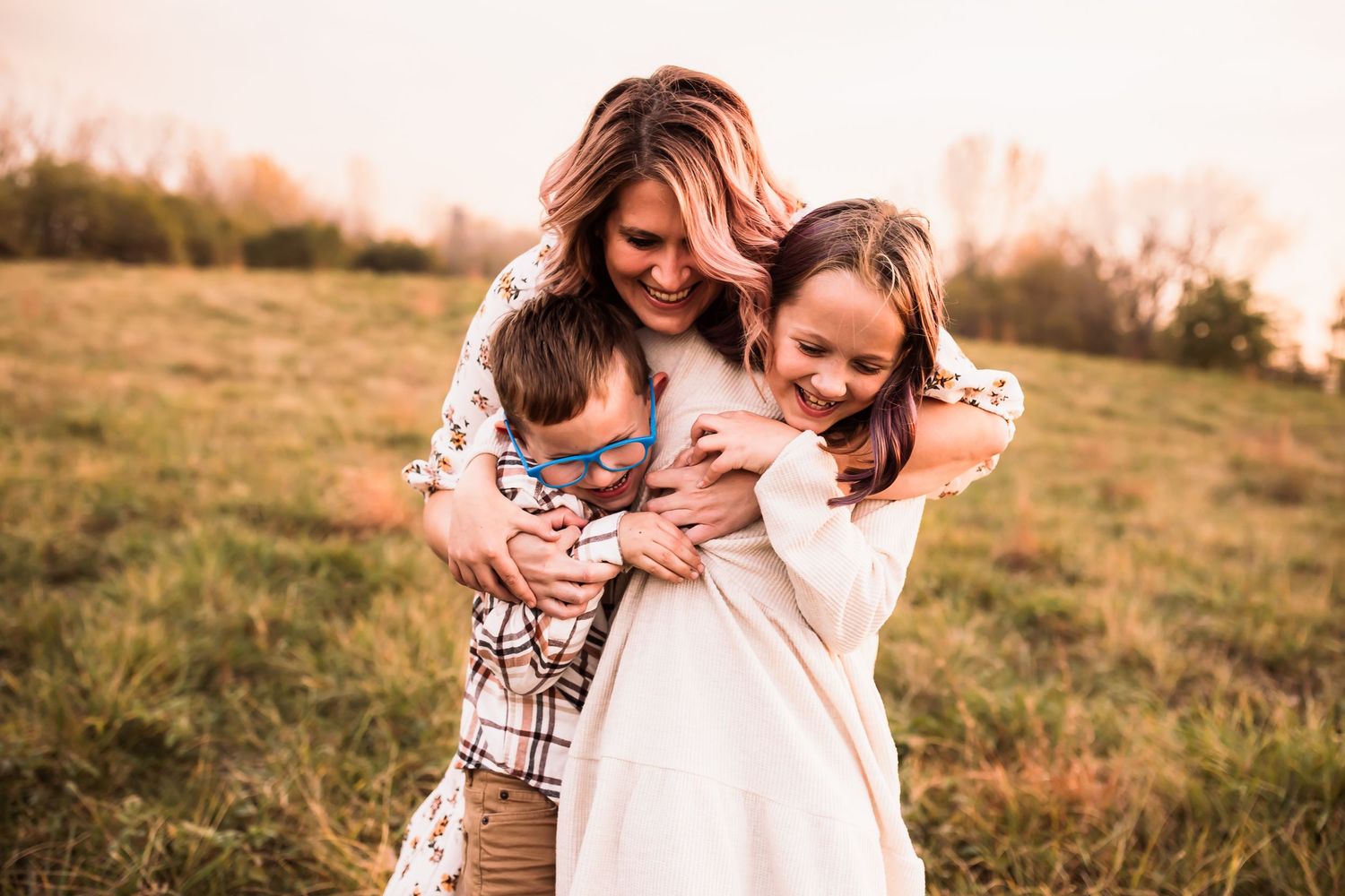 Three people embrace and laugh together in a golden sunlit field during sunset.