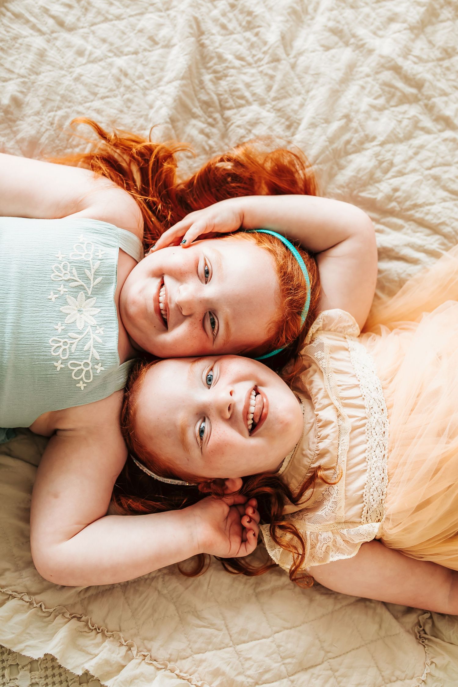 Two redheaded children wearing green and cream colored dresses lay head to head on cream colored bedding.