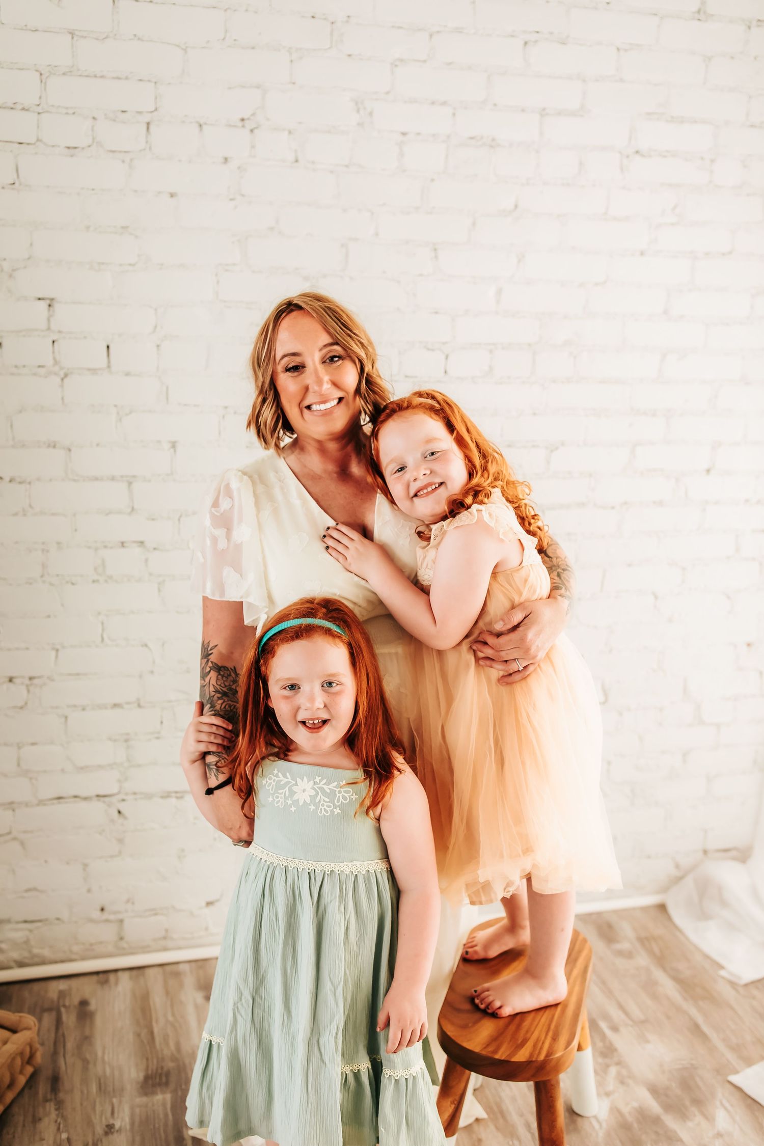 Two children in pastel dresses pose with their parent against a white brick wall in a bright studio setting.