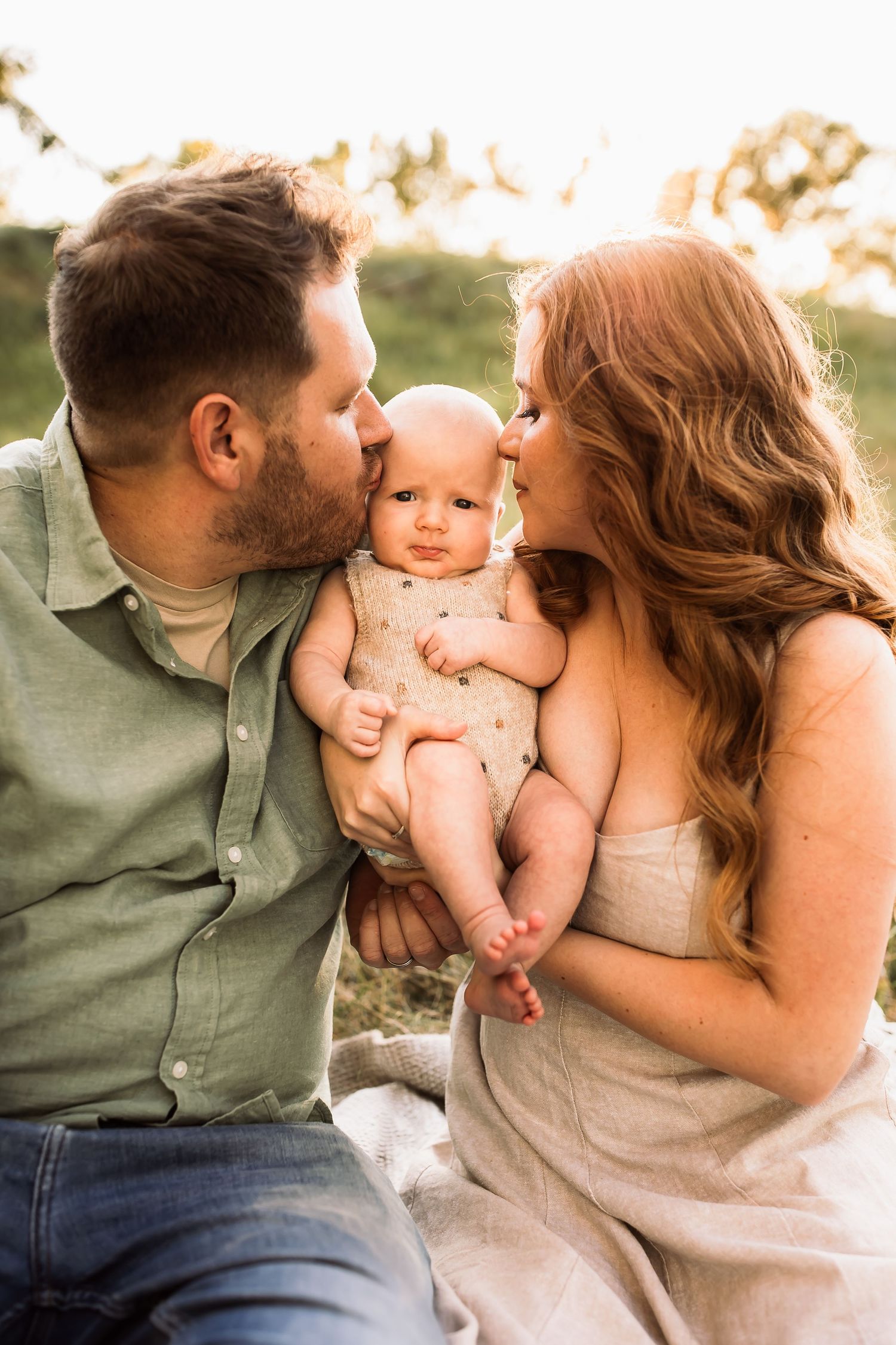 A loving family moment with parents kissing their baby during a warm outdoor photoshoot at sunset.