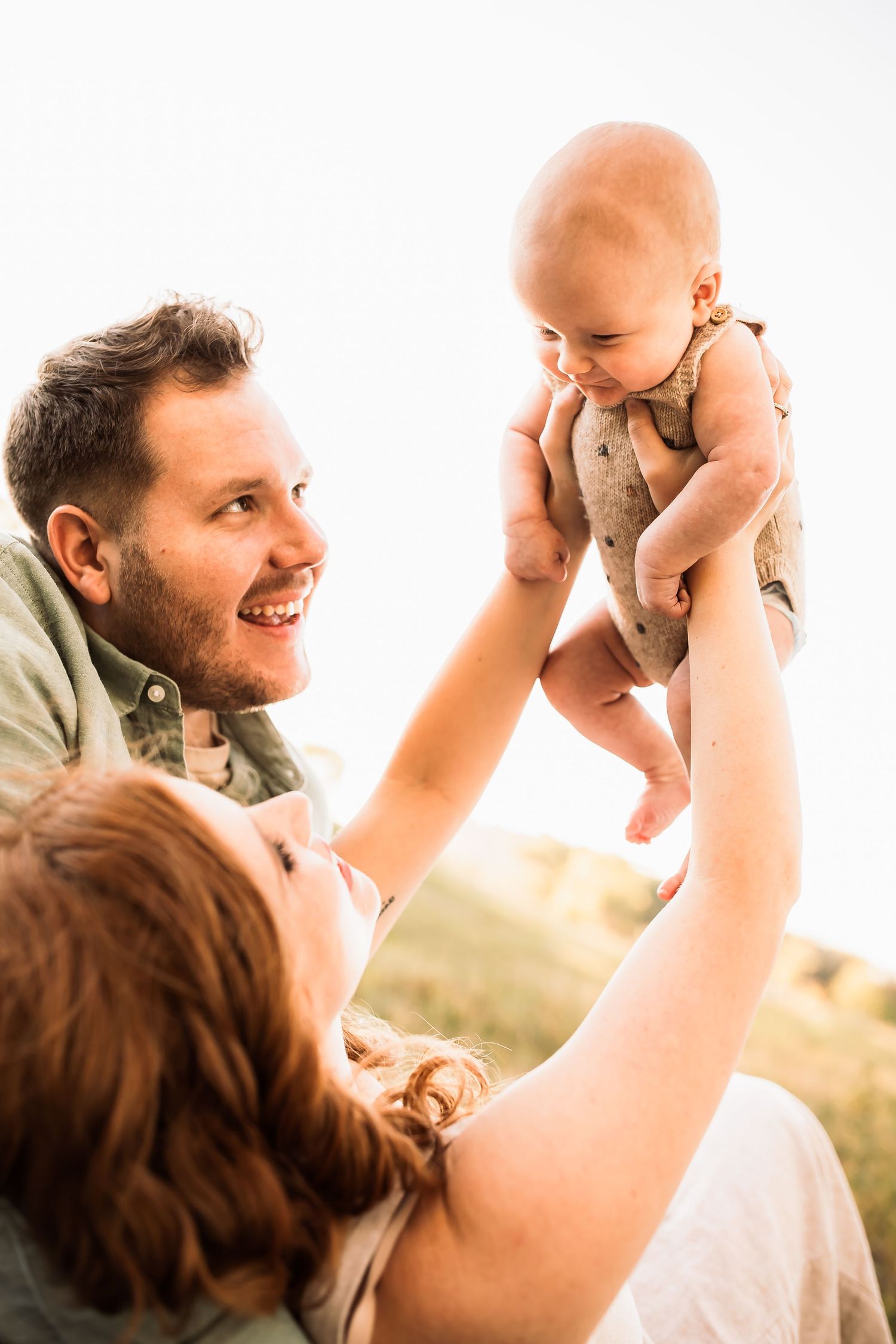 Family enjoys tender moment together outdoors lifting baby in sunlit setting.
