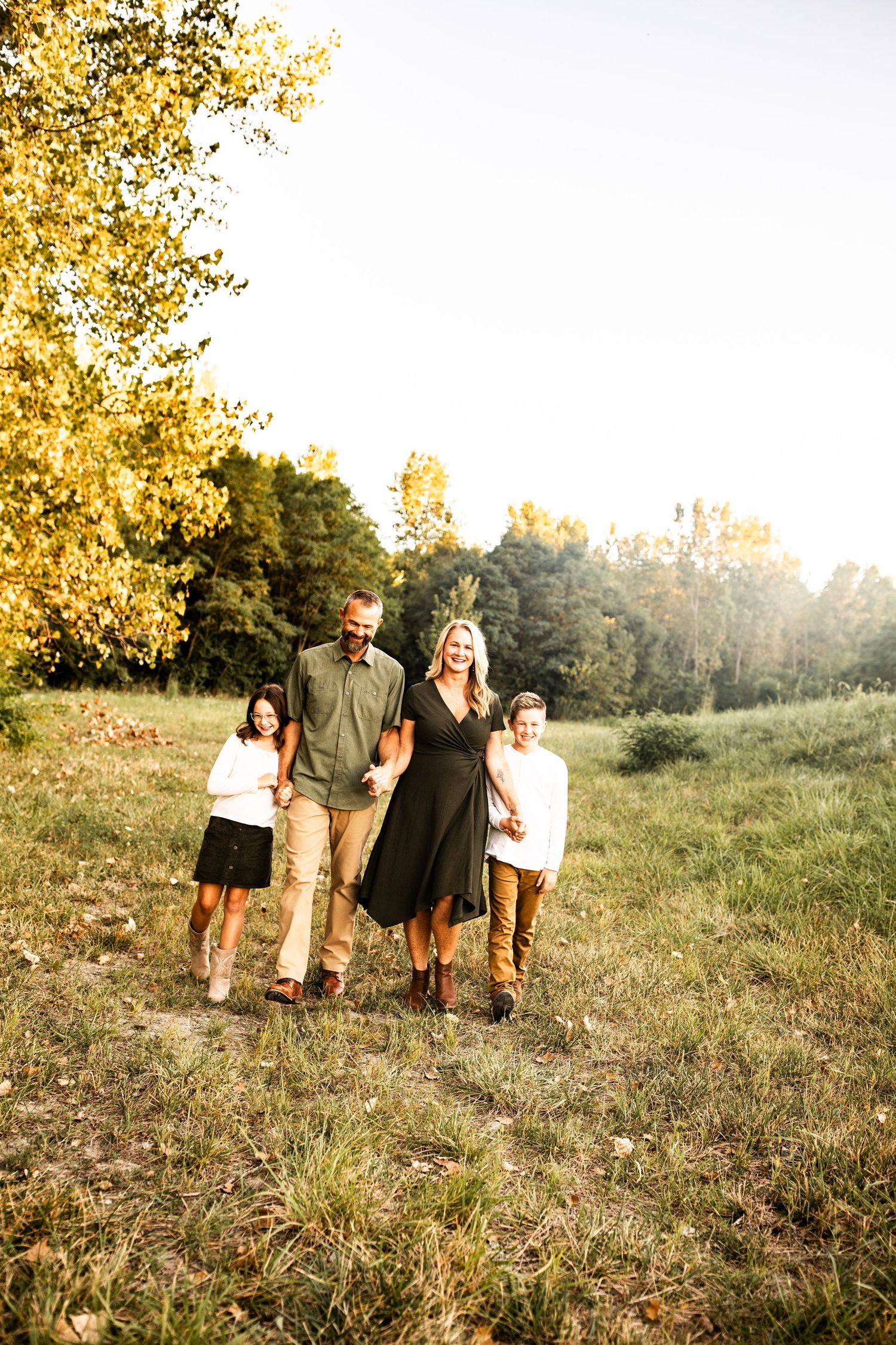 A family of four stands together in a sunlit meadow during golden hour with autumn trees in the background.
