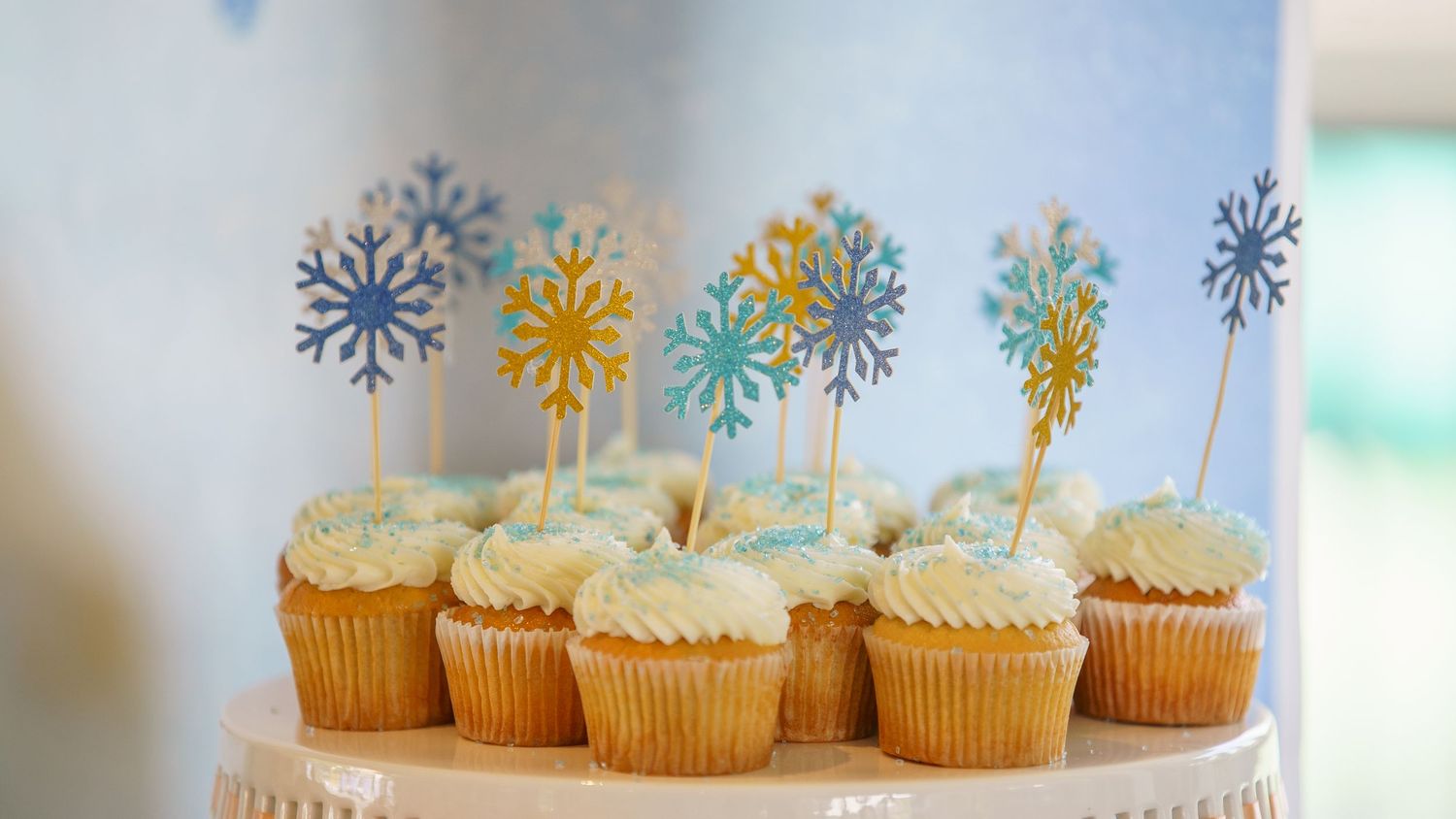 Vanilla cupcakes topped with white frosting and blue, gold, and white snowflake decorative toppers on display.