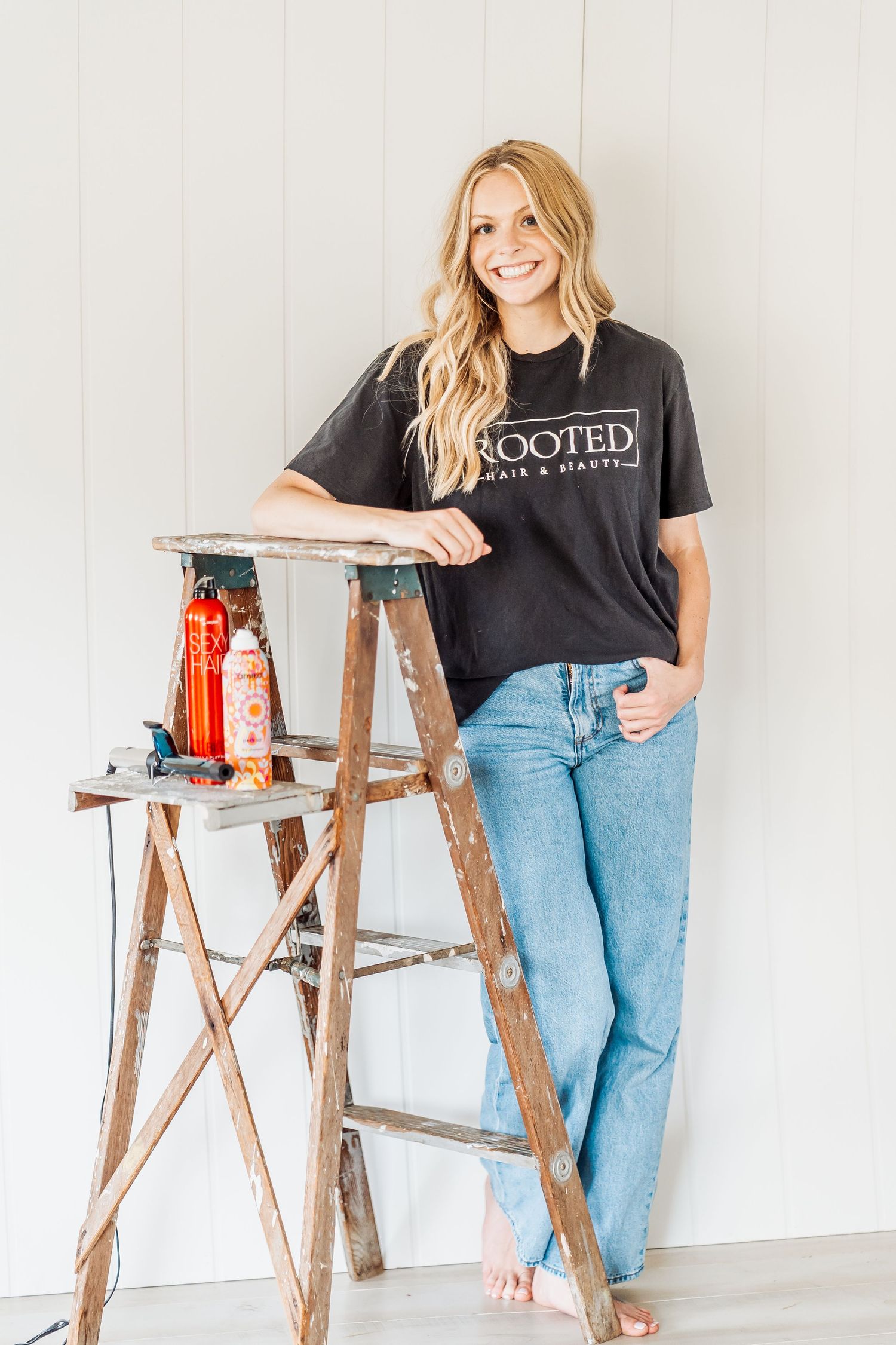 Someone wearing a black graphic t-shirt and blue jeans leans casually against a wooden ladder with decorative items nearby.