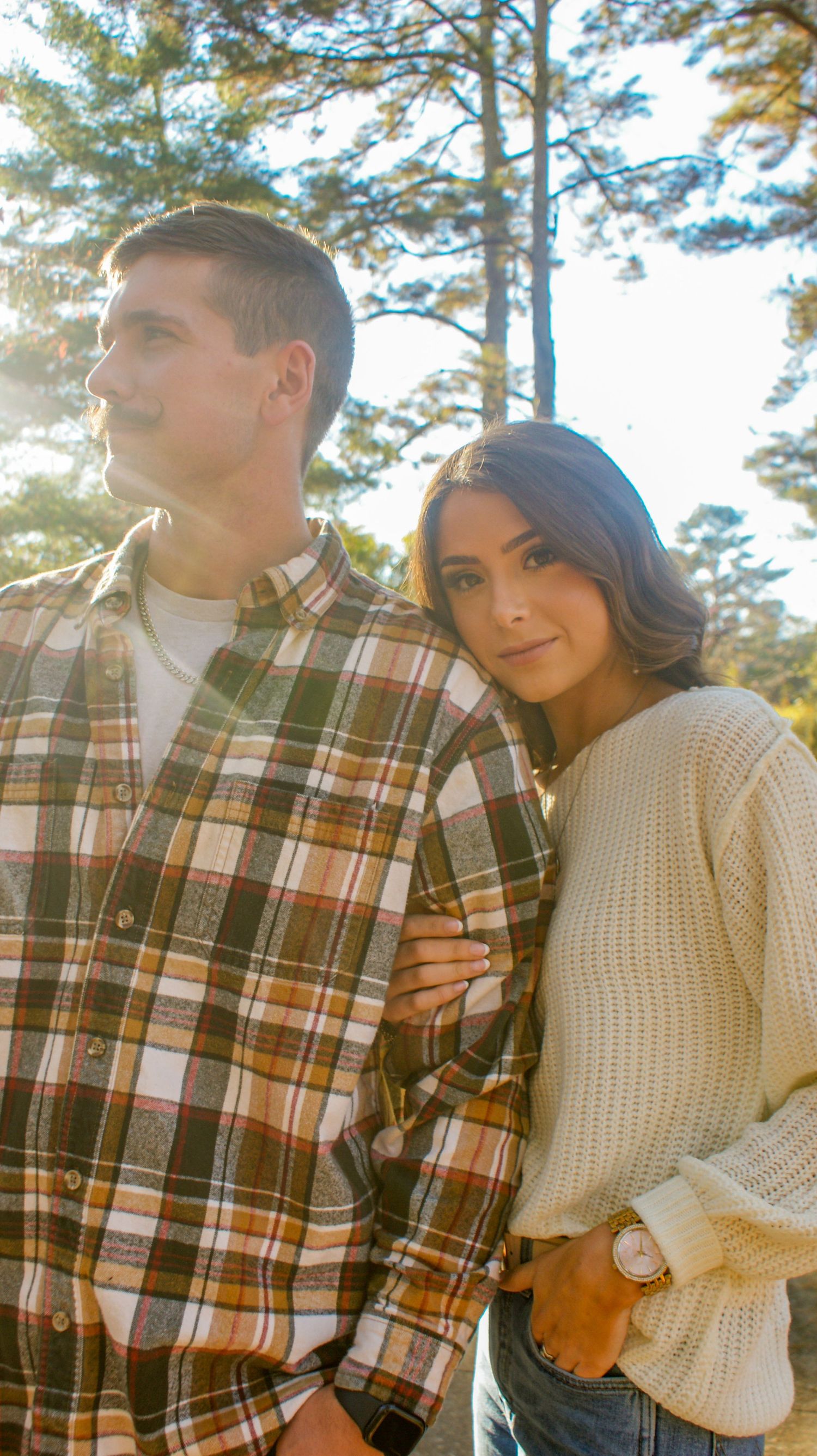 A couple poses together in casual fall attire against a backdrop of sunlit pine trees.