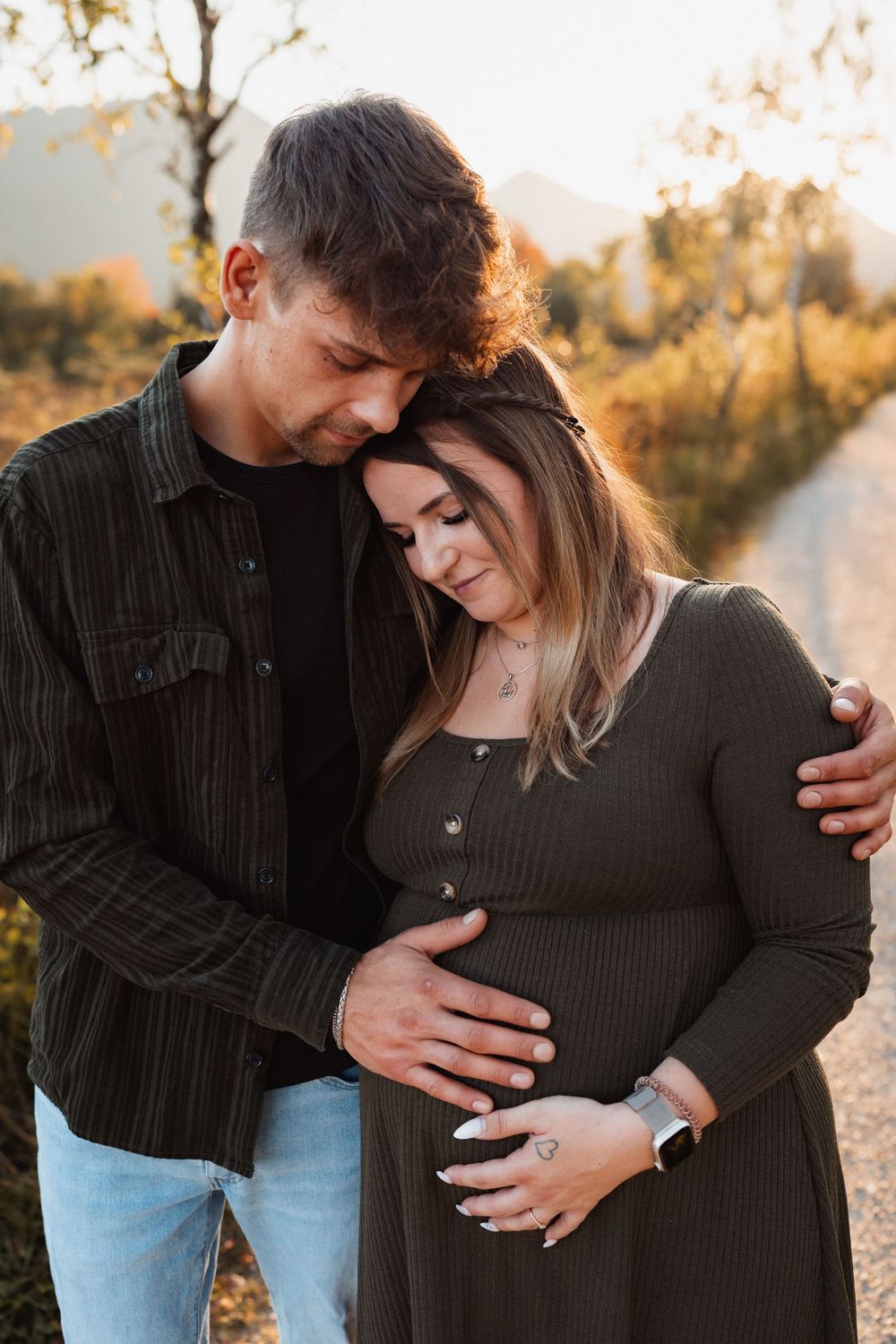 Paar hält sich im Arm, während er liebevoll den Babybauch berührt – warmes Babybauchshooting bei Sonnenuntergang