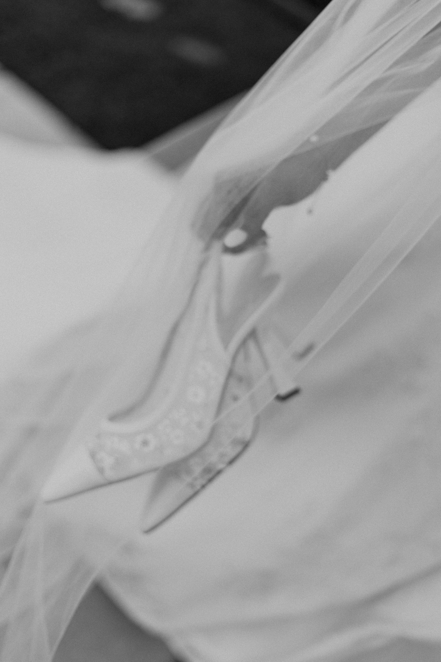 Black and white close-up of elegant lace bridal heels resting on flowing white wedding dress and veil.