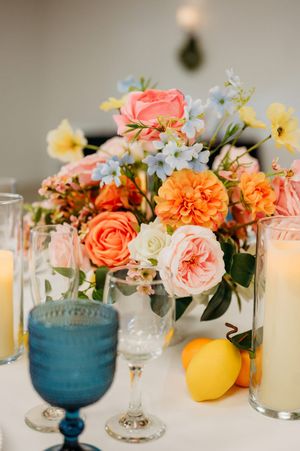 Vibrant wedding centerpiece with coral, orange, and pink flowers beside lemons and blue glassware.